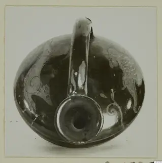 Dark glossy pottery vessel with a tall loop handle and pale painted motifs, photographed from the front.