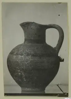 Pottery White Painted I jug with a tall neck, one loop handle and a round body, photographed against a grey backdrop.