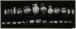 Pottery from Vouni Tomb 13: a row of jugs and juglets above a row of bowls and bowl fragments, on a dark background.