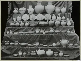 Several dozen pottery vessels arranged in tiers on a stepped stand covered with dark cloth, Lapithos Kastros tombs