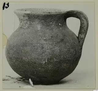 Coarse ware jug with a single handle and a short flared neck, shown in grey-brown clay against a pale background.