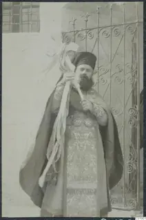 Bearded priest in a dark cap and embroidered robes holding a cross on a staff, standing before an iron gate