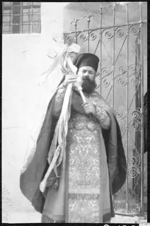 A bearded priest in embroidered vestments holding a cross, standing before an iron gate at Lapithos.