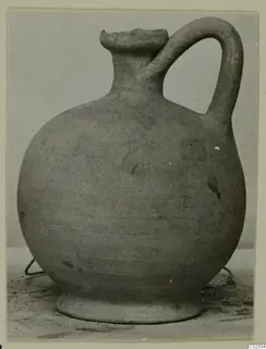 Pottery jug with a round body, a single loop handle and a short collar neck, photographed on a plain surface.