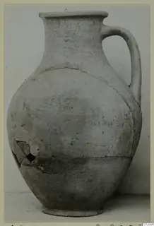 Plain pale pottery jug with a single handle, a flared rim and a cracked, repaired body, shown against a grey backdrop.