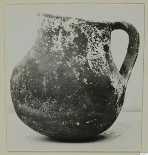 Coarse pottery jug with a single loop handle, dark surface with pale encrusted patches