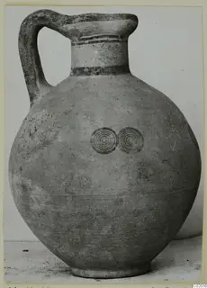 Pottery jug with a high loop handle, banded neck and two concentric circle motifs on the body.