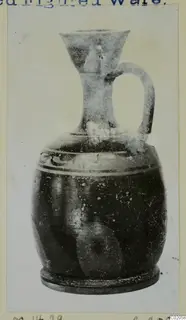 Dark jug with a funnel-shaped mouth, strap handle and pale bands around the body, shown on a mounted print.