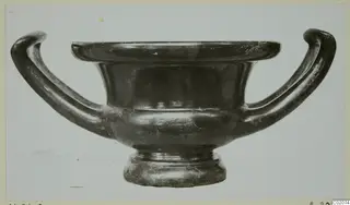Black glazed kantharos with two curving handles, photographed in side view against a pale background