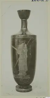Tall vessel with a flaring mouth and a painted standing figure holding a spear, photographed against a grey backdrop