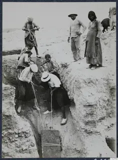 Workers haul a wooden box out of a narrow excavation trench on ropes, with two onlookers standing at the edge