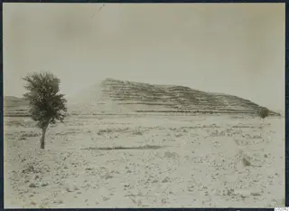 Wide view of a layered hill rising above stony ground, with a tree at left and a smaller tree to the right, at Idalion