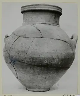 Large plain pottery vessel with a wide rim and two handles, cracked and pieced back together, against a pale background.
