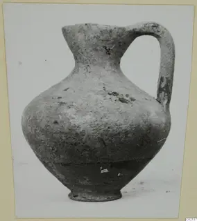 Pottery juglet with a single strap handle and a flaring neck, shown on a pale surface
