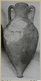 Two-handled pottery jar with a narrow neck and pointed base, pale surface with darker patches and cracks, against a grey backdrop.