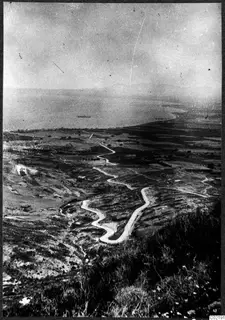 Winding road descending from a hillside toward a coastal plain and the sea, with a ship offshore.