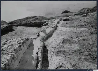 Excavated rock-cut channel and low walls at the palace at Vouni, with hills and a pale open view beyond.
