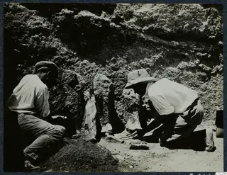 Two men kneeling in a trench uncovering a standing stone statue at Vouni