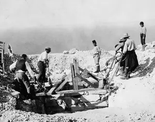Men and a boy work with ropes beside a stone-lined cistern at Vouni, with the sea visible behind.
