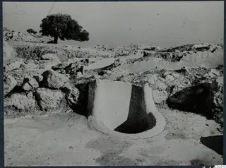 Excavated area at Vouni with a round plastered opening in the foreground, rough stones and a tree behind.