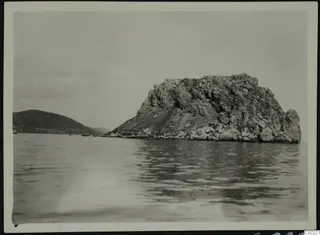 Rocky islet rising from the sea at Petra tou Limniti, with a low hill on the left.