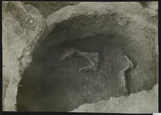 Skeleton lying in an excavated pit at Ayios Iakovos, with bones partly uncovered in the dark soil.