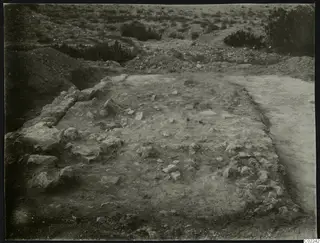 Excavated area at Ayios Iakovos with exposed stone walls, scattered stones and low scrub beyond.