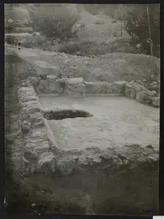 Excavated area at Ayios Iakovos with a low stone wall enclosing a pale floor and a round pit