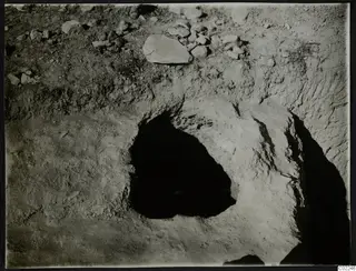Excavation at Ayios Iakovos with a dark opening in the soil and scattered stones on the surface.