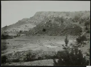 Rocky hillside at Ayios Iakovos with pale excavation spoil heaps, a tall tree and a small group of people at the right edge.