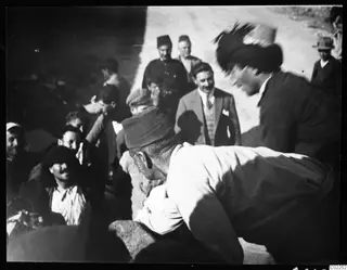 Crowd of men on a dirt road, one bending over in the foreground while others look on