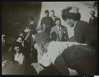Men gathered on a dusty road, one bending over a pale rounded object, others standing and seated around him in caps and fezzes
