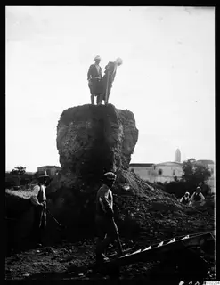 Two men stand atop a large earthen mound being torn down at Kition, with four workers below
