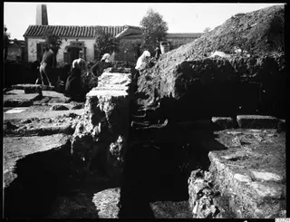 Excavation at Kition with stone walls in trenches, workers among the stonework and a whitewashed building with a tiled roof behind