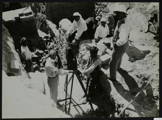 Man at a tripod-mounted camera in a trench at Kition, with workers around him among cut stone blocks