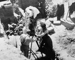 Man at a tripod-mounted camera in an excavation trench, with workers and heaped stones and objects behind him.