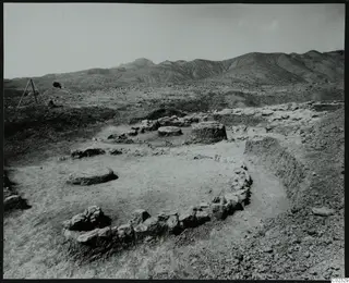Excavated house foundations with low stone walls and round stone bases on a bare floor, with hills behind, at Kythrea