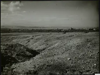Stony hillside at Kythrea with a few people, a gully on the left and a village on the plain below distant hills