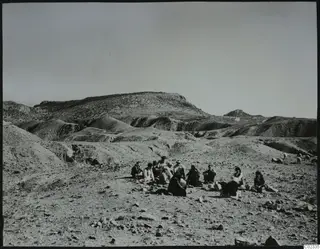 People seated on stony ground below eroded hills at Kythrea, with a flock of sheep or goats at the right edge.