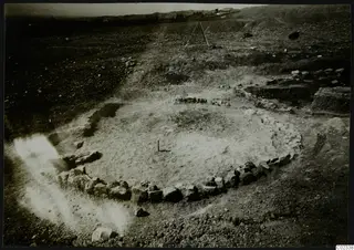 Excavated stone wall foundation at Kythrea, curved around a cleared earth floor, with hills on the horizon