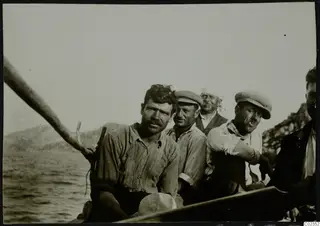 Petra tou Limniti: five men seated in a wooden boat on the water, with hills and a rocky shore behind them.
