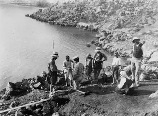 Workers at an excavation on the rocky shore at Petra tou Limniti, with a kneeling woman in a headscarf and a man in a straw hat.