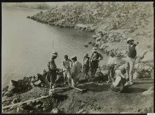 Workers and a woman in headscarf at an excavation on a rocky shoreline at Petra tou Limniti, with a cord stretched across the dig.