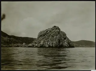 Rocky headland rising from the sea, seen from the water, with hills behind it.
