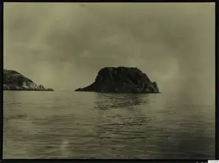 Rocky islet rising from calm sea, with a second rocky headland at the left edge under a grey sky.