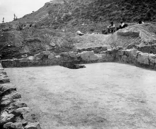 Cement floor of an excavated room at Ayios Iakovos, bounded by stone walls, with three people seated on the spoil heap beyond