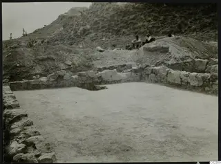 Cement floor at Ayios Iakovos enclosed by stone walls, with people seated at the edge of the excavation