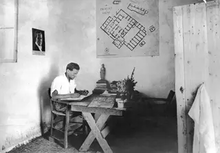 Alfred Westholm writing at a desk in a room at Vouni, with a plan of the palace on the wall behind him.