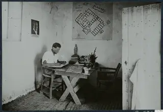 Alfred Westholm writing at a wooden table in a room at Vouni, with a plan of the palace on the wall behind him.