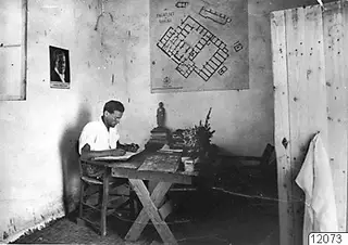 Alfred Westholm seated at a wooden desk in Vouni, with a plan of the palace on the wall behind him